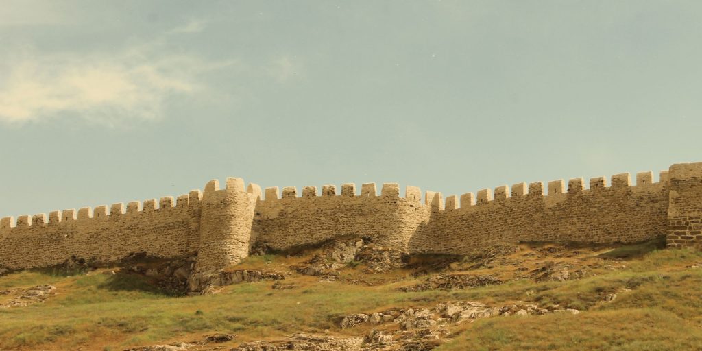 A historic stone fortress wall stretches along a grassy hill under a clear blue sky.